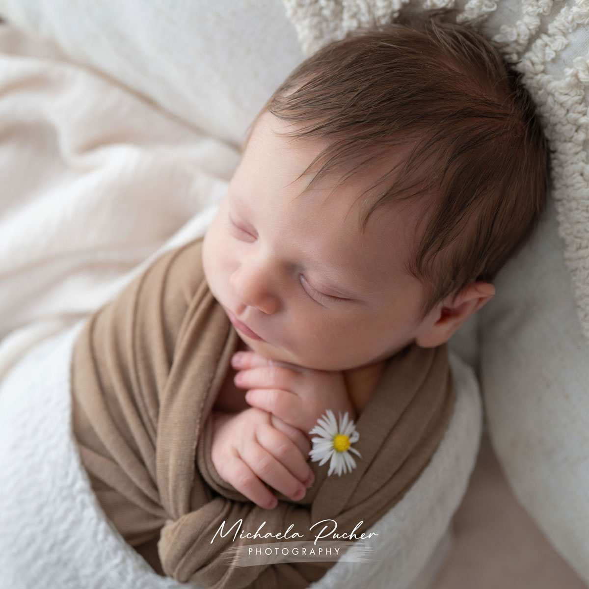 Schlafendes Neugeborenes, eingewickelt in braune und weiße Tücher, hält eine kleine Gänseblume zwischen den Fingern.